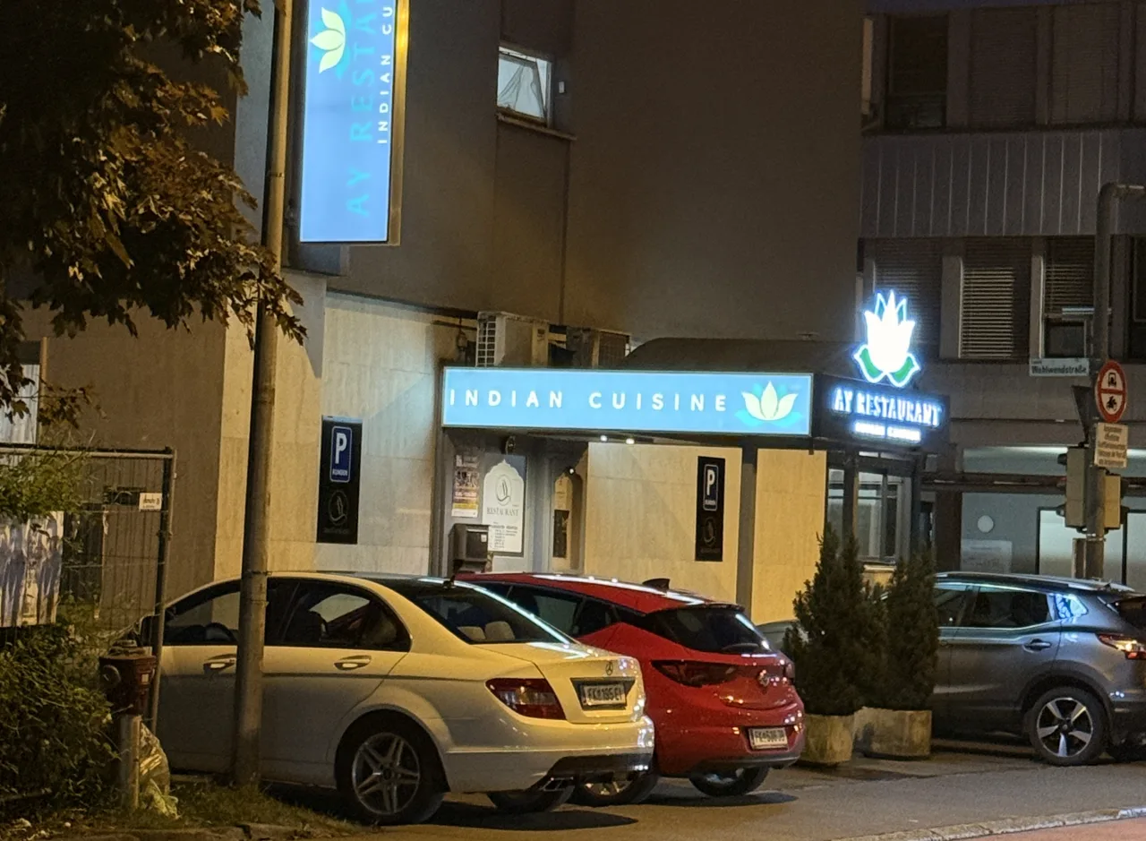 Dieselben Schilder des AY Restaurant bei Nacht beleuchtet: Vordachschild, Ausleger Indian Cuisine und der Eingangsbereich
