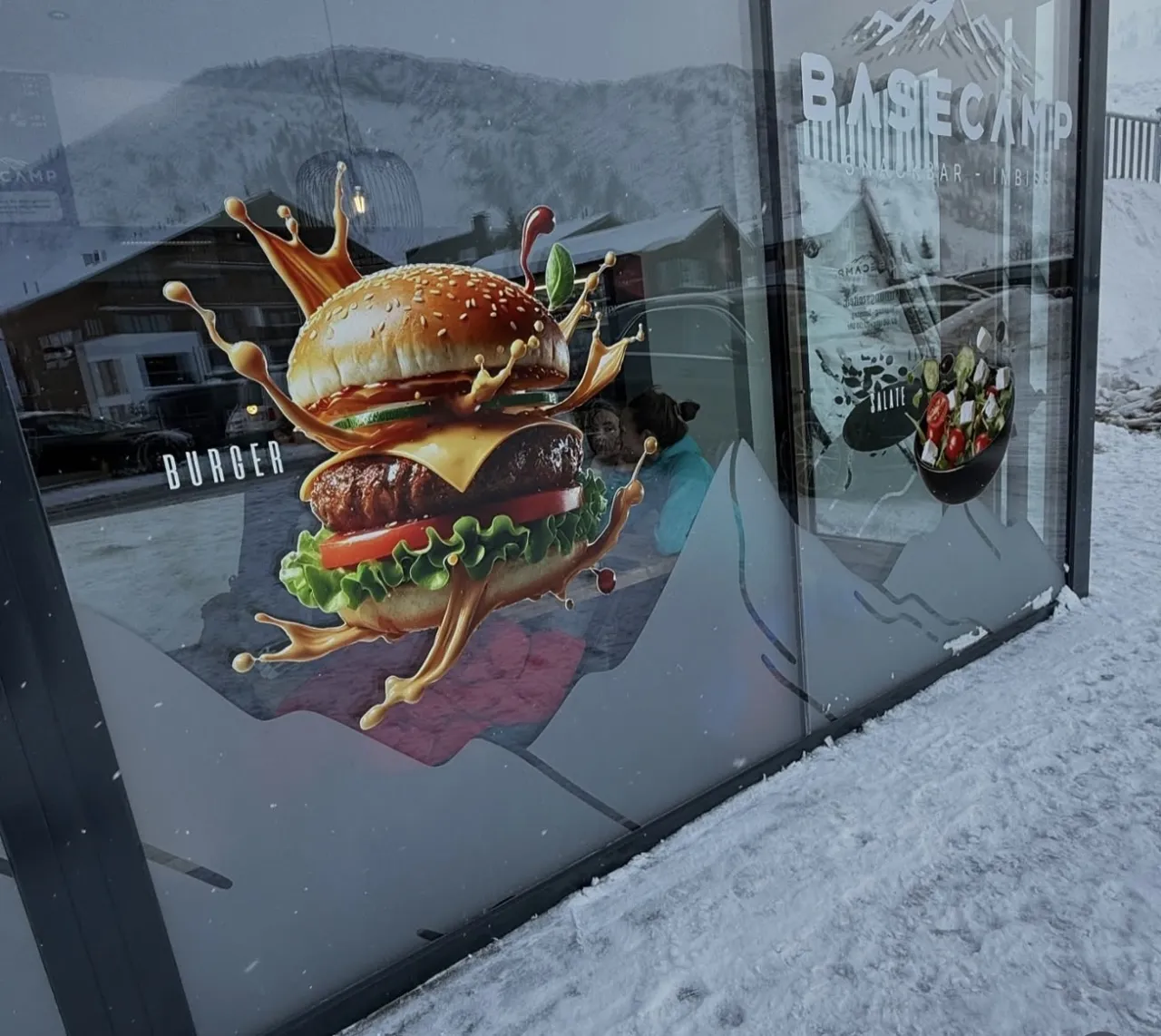 Schaufensterfolierung der Basecamp Snackbar in Warth mit Burger-Motiv und Bergsilhouette im Schnee