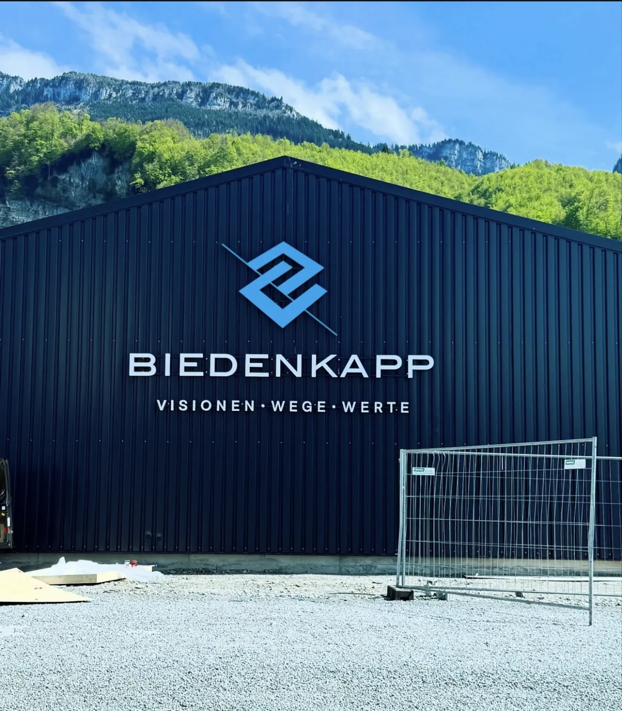 Große Hallenfassade mit 3D-Logo und Schriftzug Biedenkapp vor bewaldetem Berghang