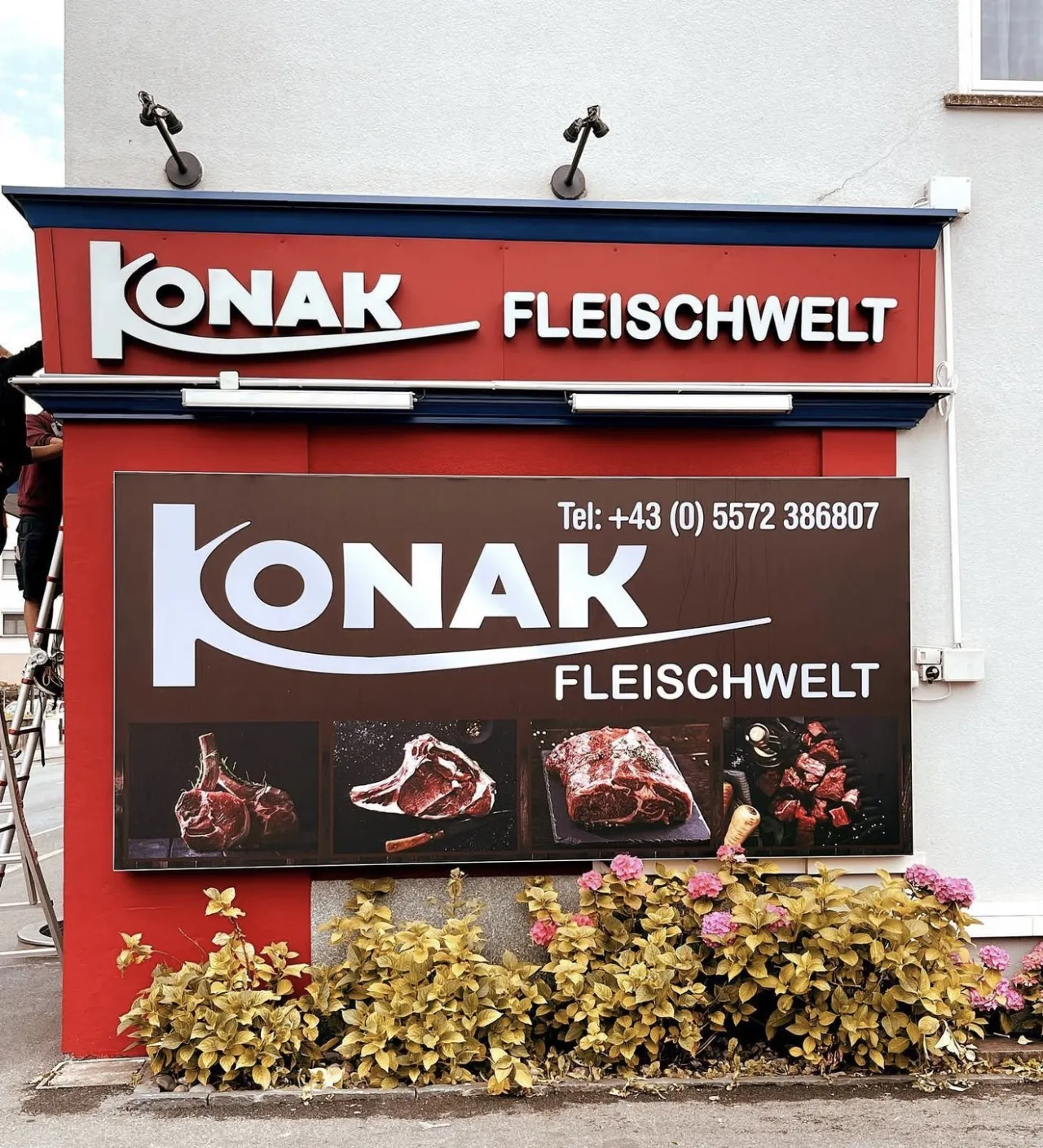 Großes bedrucktes Werbeschild der Konak Fleischwelt mit Fleischmotiven, daneben eine Leiter während der Montage