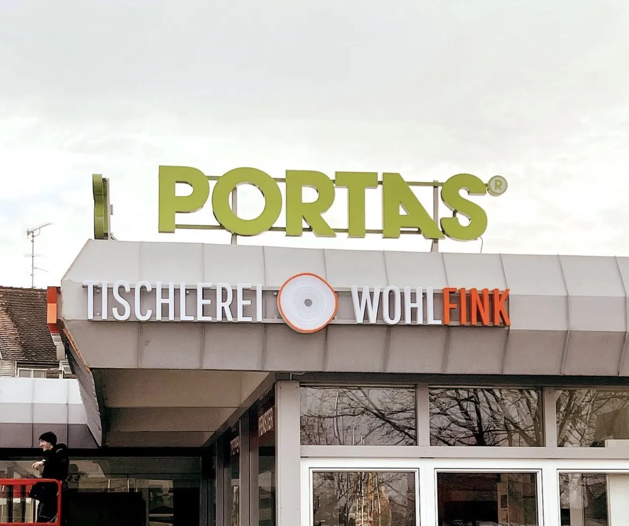 Dachschriftzug Portas über dem Fassadenband Tischlerei Wohlfink, daneben ein Monteur auf einer Hebebühne