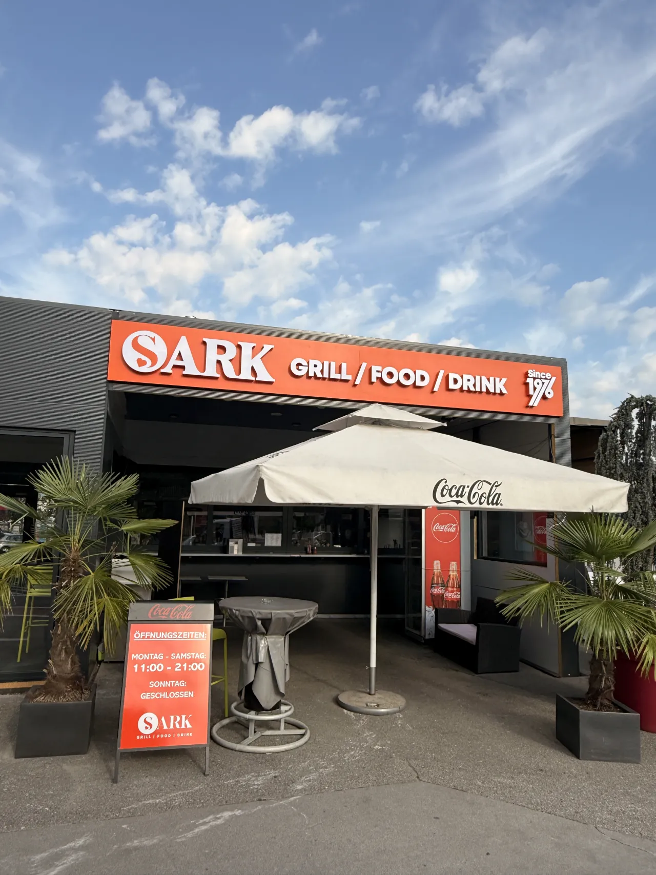 Orangefarbenes Fassadenschild von SARK Grill Food Drink mit weißen 3D-Buchstaben vor blauem Himmel
