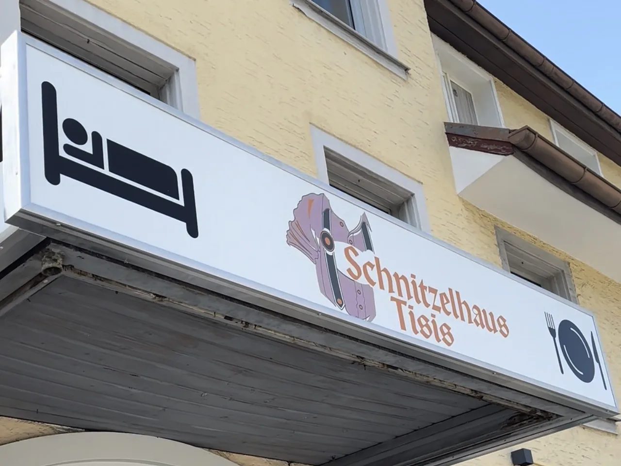 Weißer Leuchtkasten über dem Eingang des Schnitzelhaus Tisis in Feldkirch mit Bett-Piktogramm, Logo und Teller-Symbol